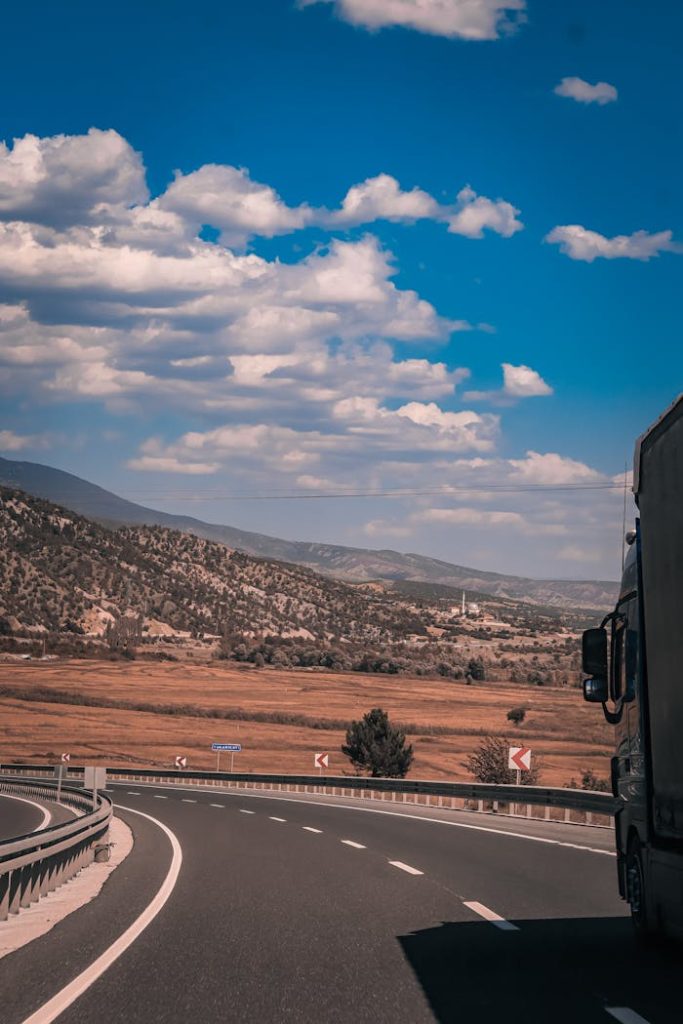 A large truck travels on a winding countryside highway beneath a clear blue sky with scattered clouds.